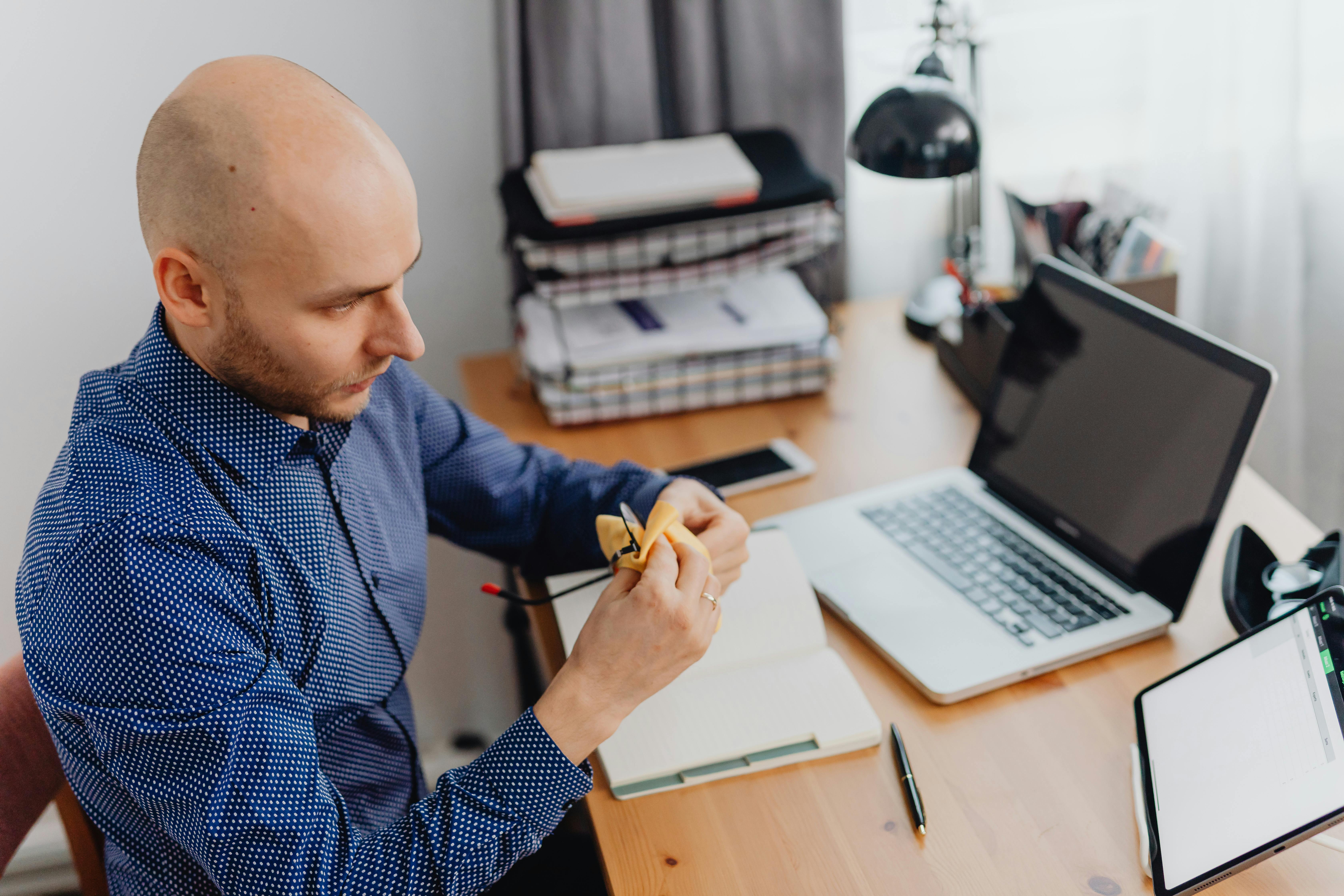 Bald man in a home office setting, reviewing documents with a laptop and tablet.