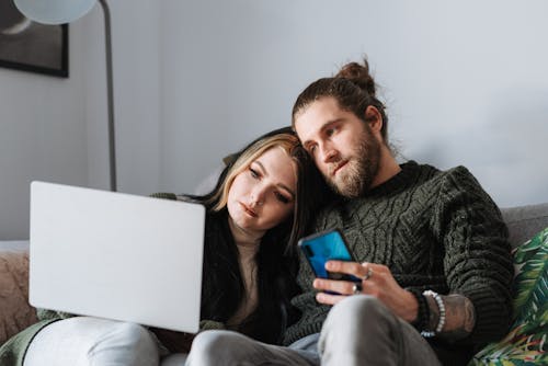 Young hipster man with cellphone near female partner surfing internet on portable computer in house room