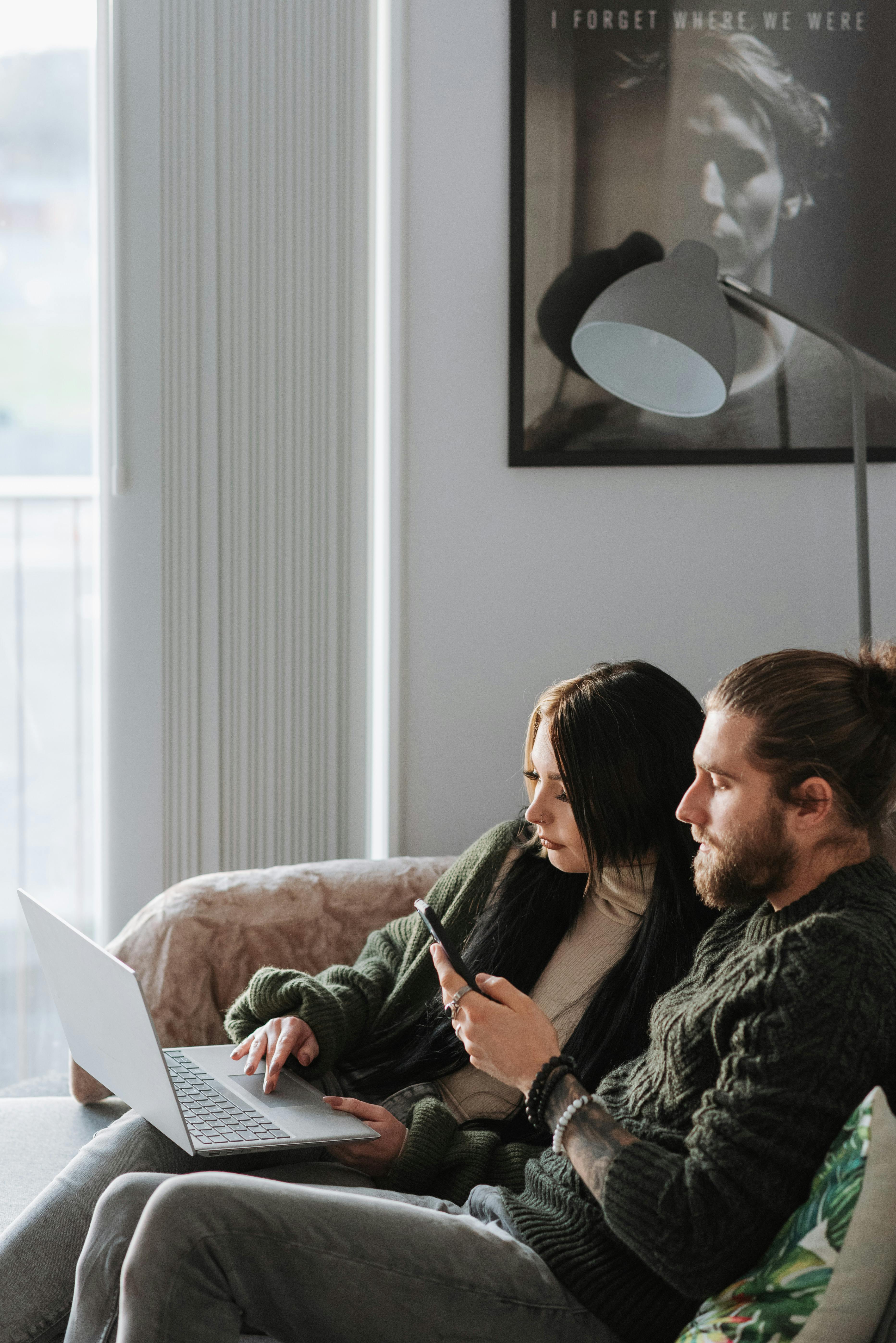 man chatting on smartphone near girlfriend with laptop on couch