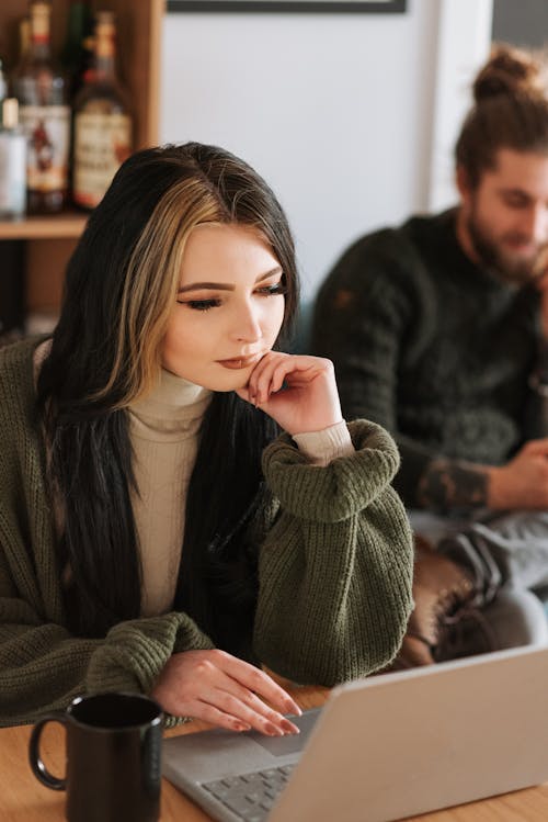 Young female touching chin while browsing internet on portable computer at table with hot drink against crop boyfriend in house