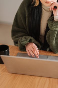From above of crop anonymous female remote employee working on portable computer near mug of hot beverage while touching chin in house