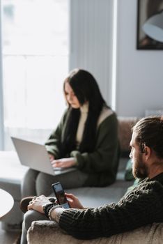 Crop young unshaven male browsing internet on cellphone near female partner typing on netbook on sofa in living room