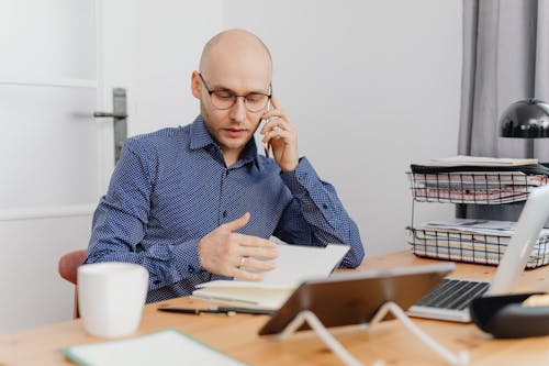 Man in Blue and White Polka Dots Dress Shirt Using Cellphone While Looking At His Notebook