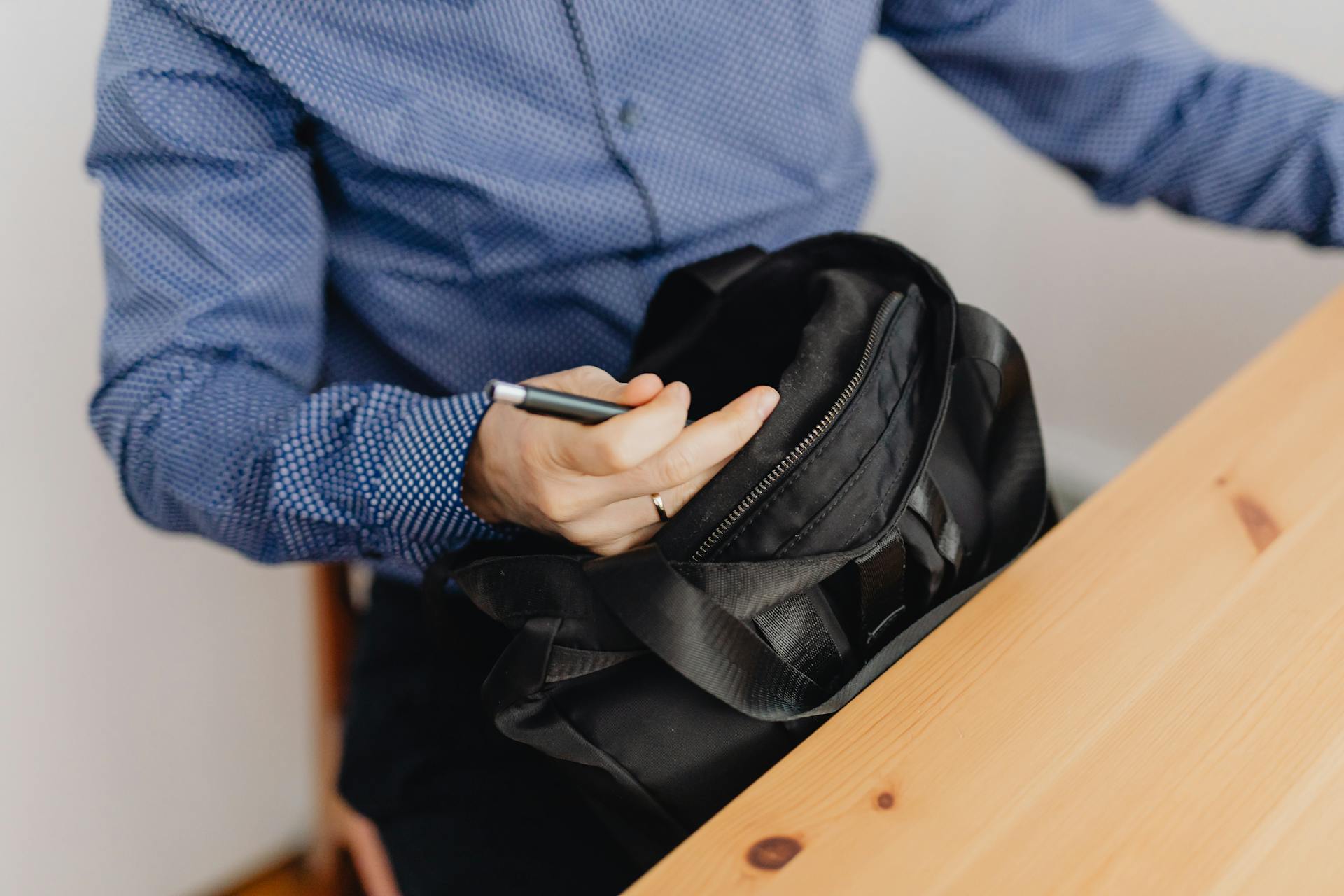 Close-up of a businessman opening a black leather bag on a wooden desk, holding a pen.