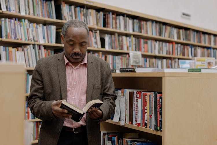 Man Standing By A Bookshelf In A Library And Reading A Book 