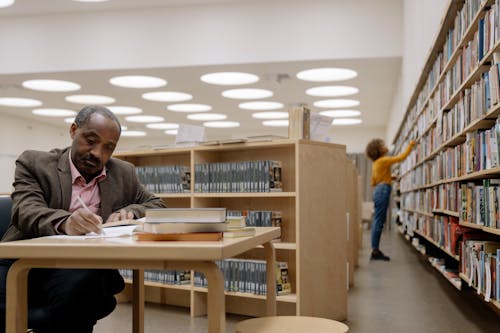 Free Man in Brown Suit Jacket Writing on Table Inside the Library Stock Photo