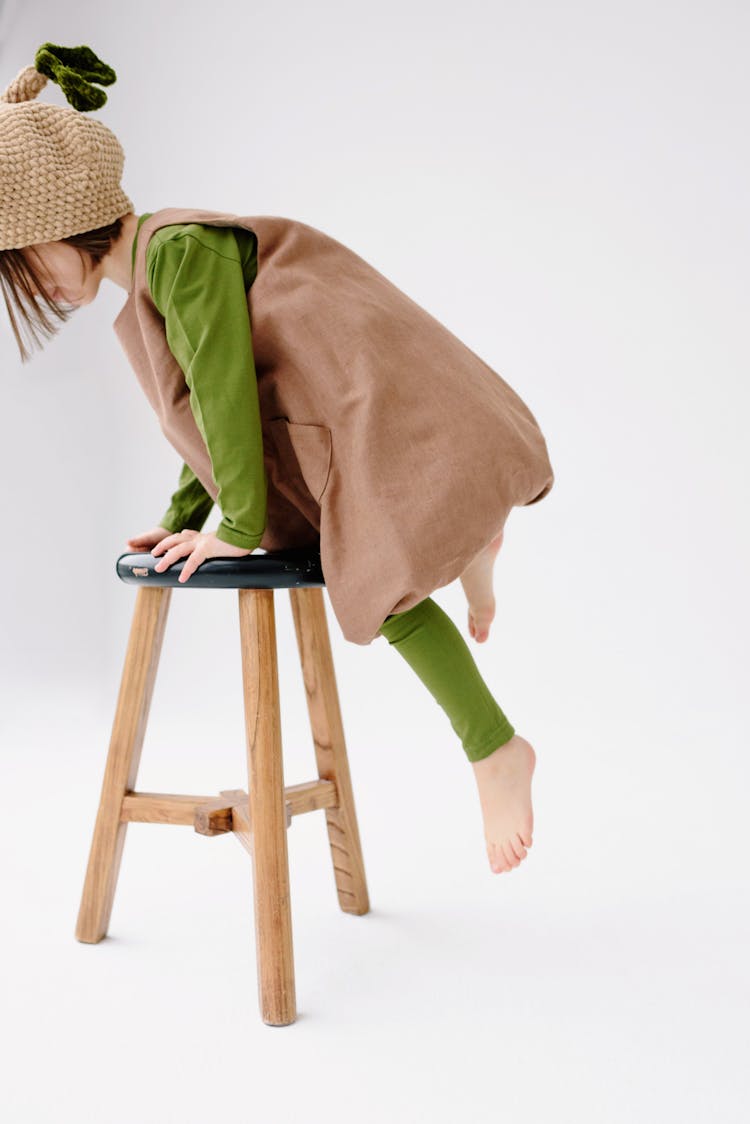 Girl In Brown Loose Romper With Green Leggings And Knit Hat Climbing Onto A Wooden Chair