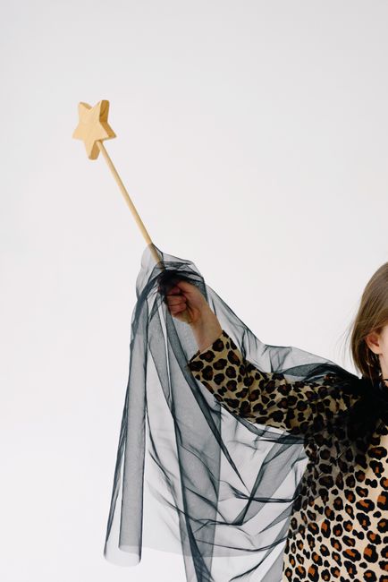 A child wearing a leopard print costume holds a wooden star wand against a plain backdrop.