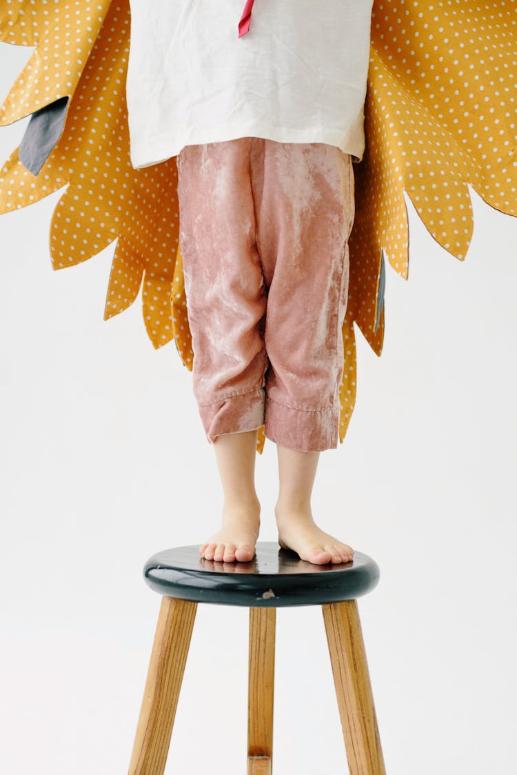 Low Section Of A Child In Costume With Yellow Wings And Pink Velvet Trousers Standing On A Chair