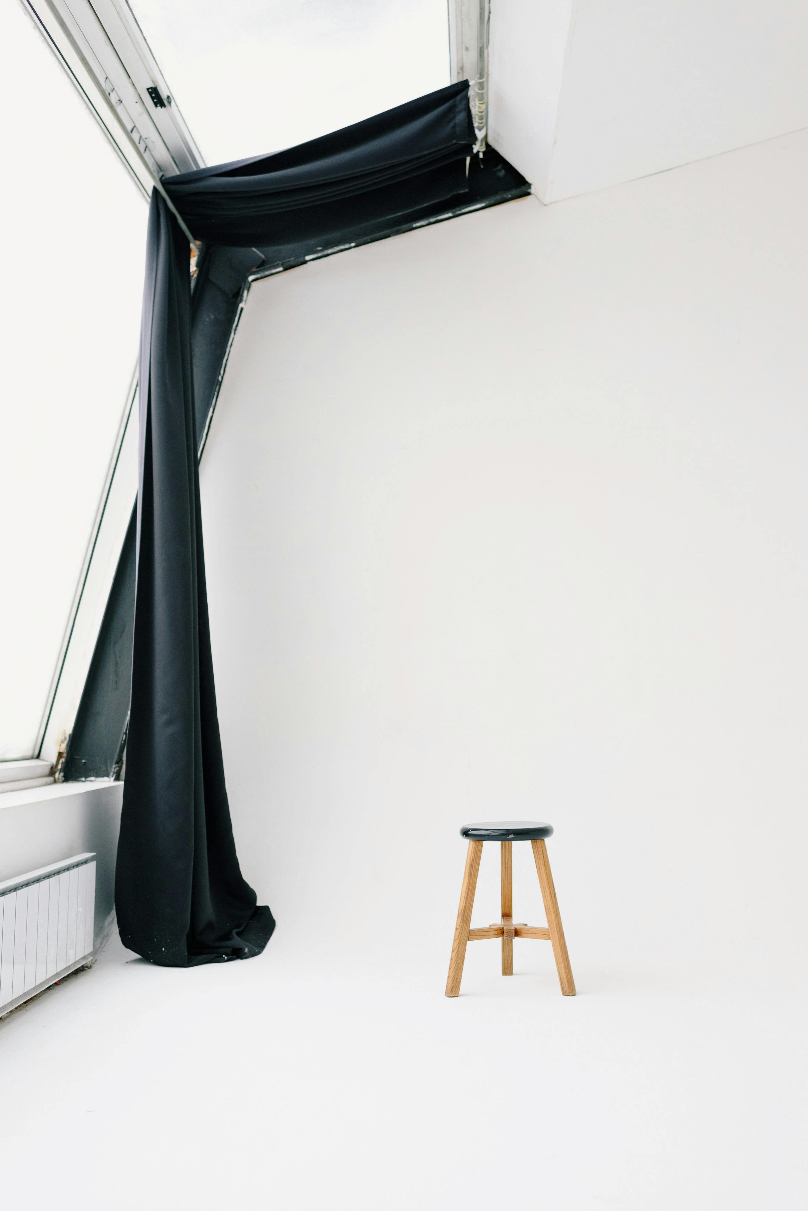 A simple white room interior featuring a single wooden stool and a draped black curtain.