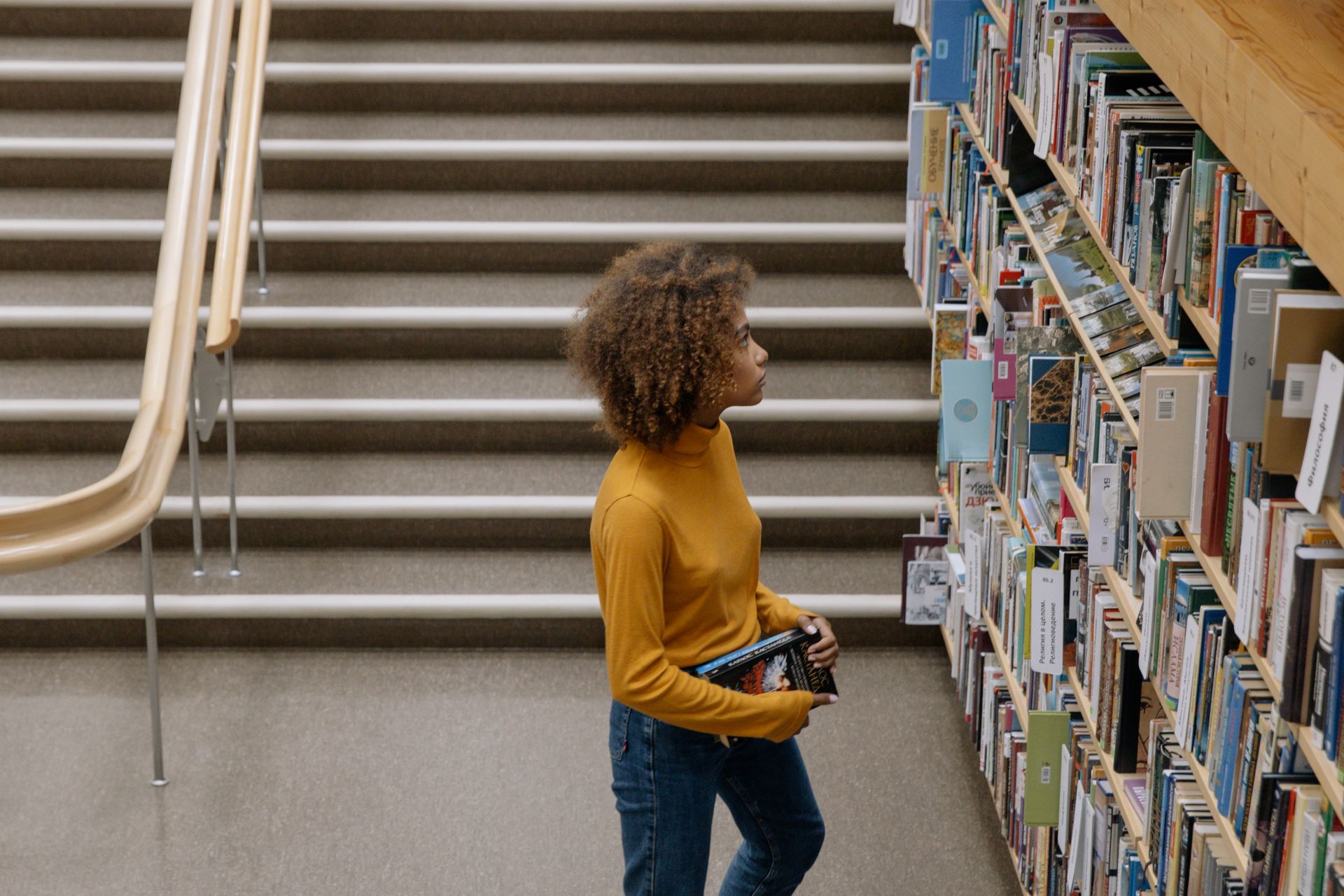 Young woman in yellow sweater browsing books in a library with shelves and stairs.