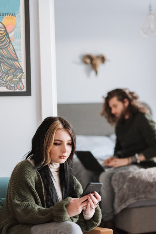 Young attentive female chatting on cellphone against male partner sitting on bed in house