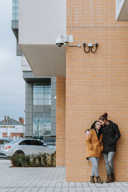 Young hipster man embracing gently female partner while leaning on brick building wall in city