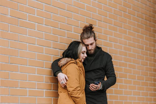 Young unshaven man embracing female partner while surfing internet on cellphone near wall in daytime