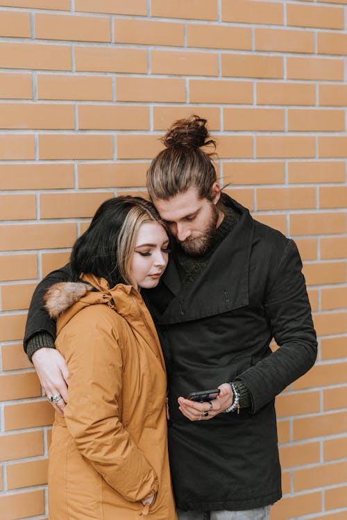 Calm young loving couple in warm outerwear hugging and using smartphone while standing on street near brick building