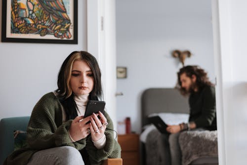 Thoughtful young informal female millennial in casual clothes sitting in armchair and messaging on smartphone during weekend at home with bearded boyfriend working remotely on laptop