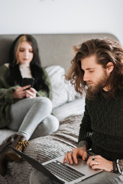 Serious young couple using laptop and smartphone sitting on bed at home