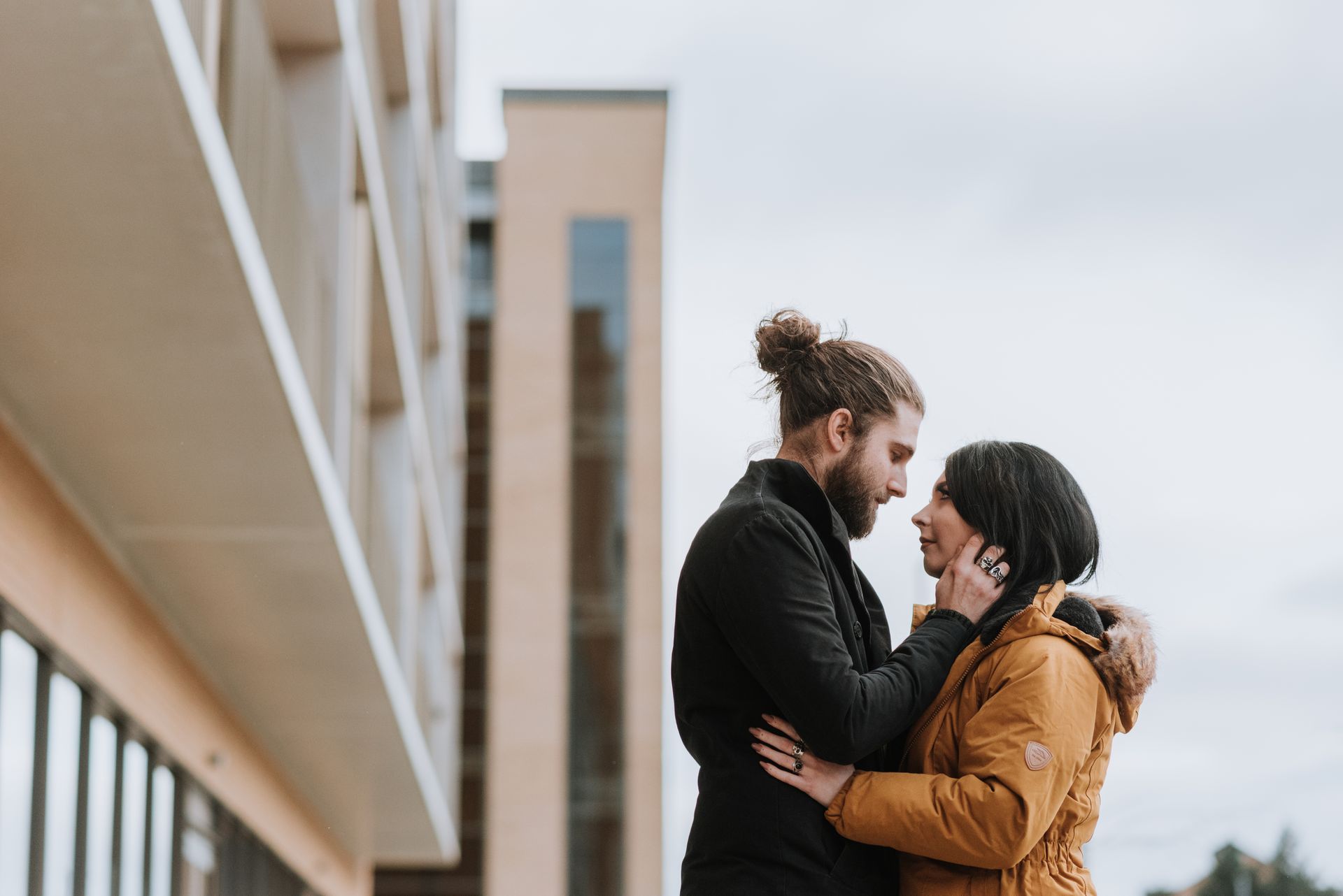 Side view of enamored young bearded man in outerwear touching face of girlfriend and looking at each other near modern building on city street