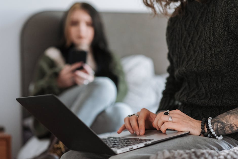 Crop faceless young male remote worker using laptop while sitting on bed at home near anonymous girlfriend browsing mobile phone