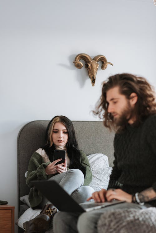 Serious young female in warm clothes messaging on mobile phone while resting on bed near concentrated boyfriend working remotely on laptop