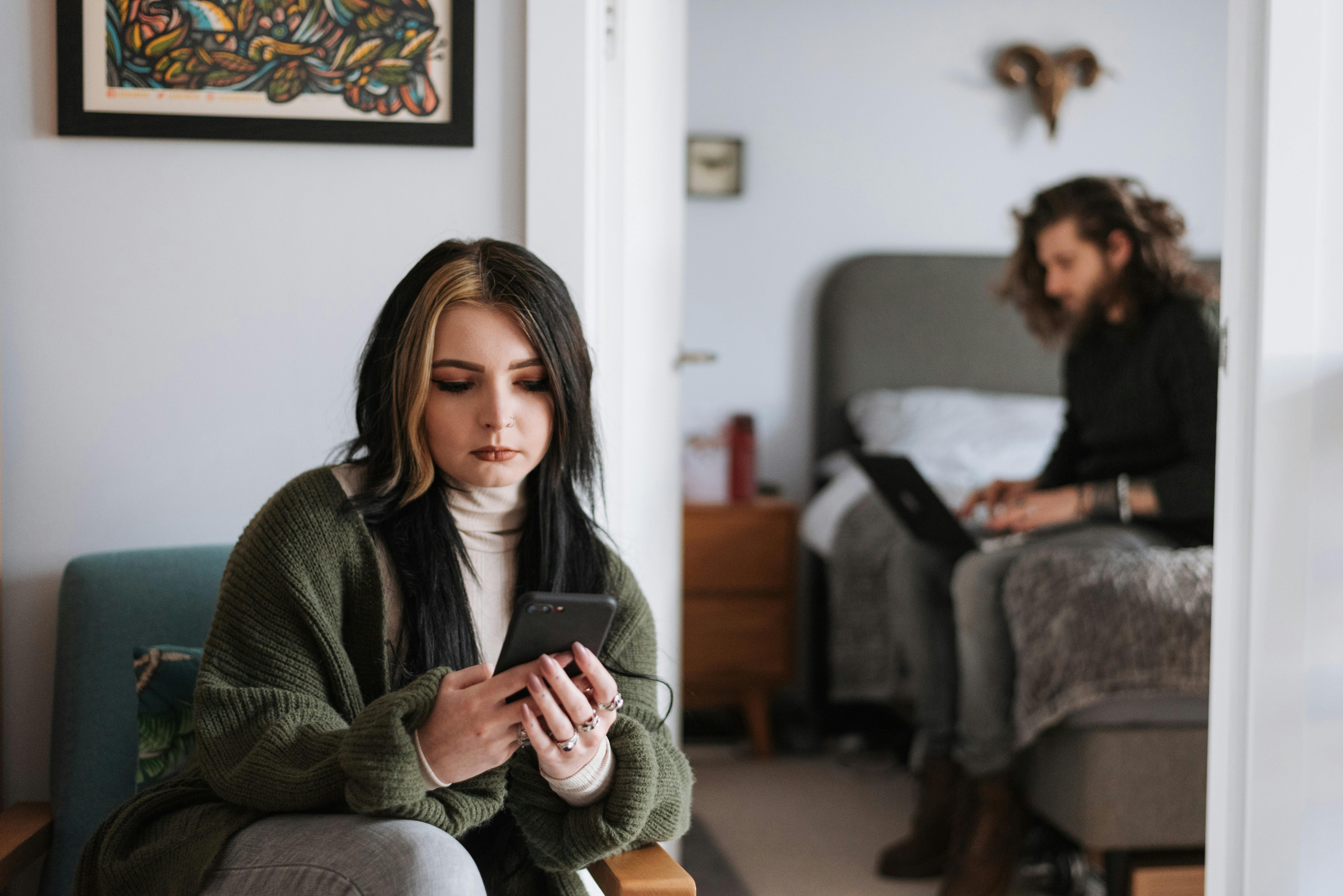 pensive woman using mobile phone at home near boyfriend working remotely on laptop