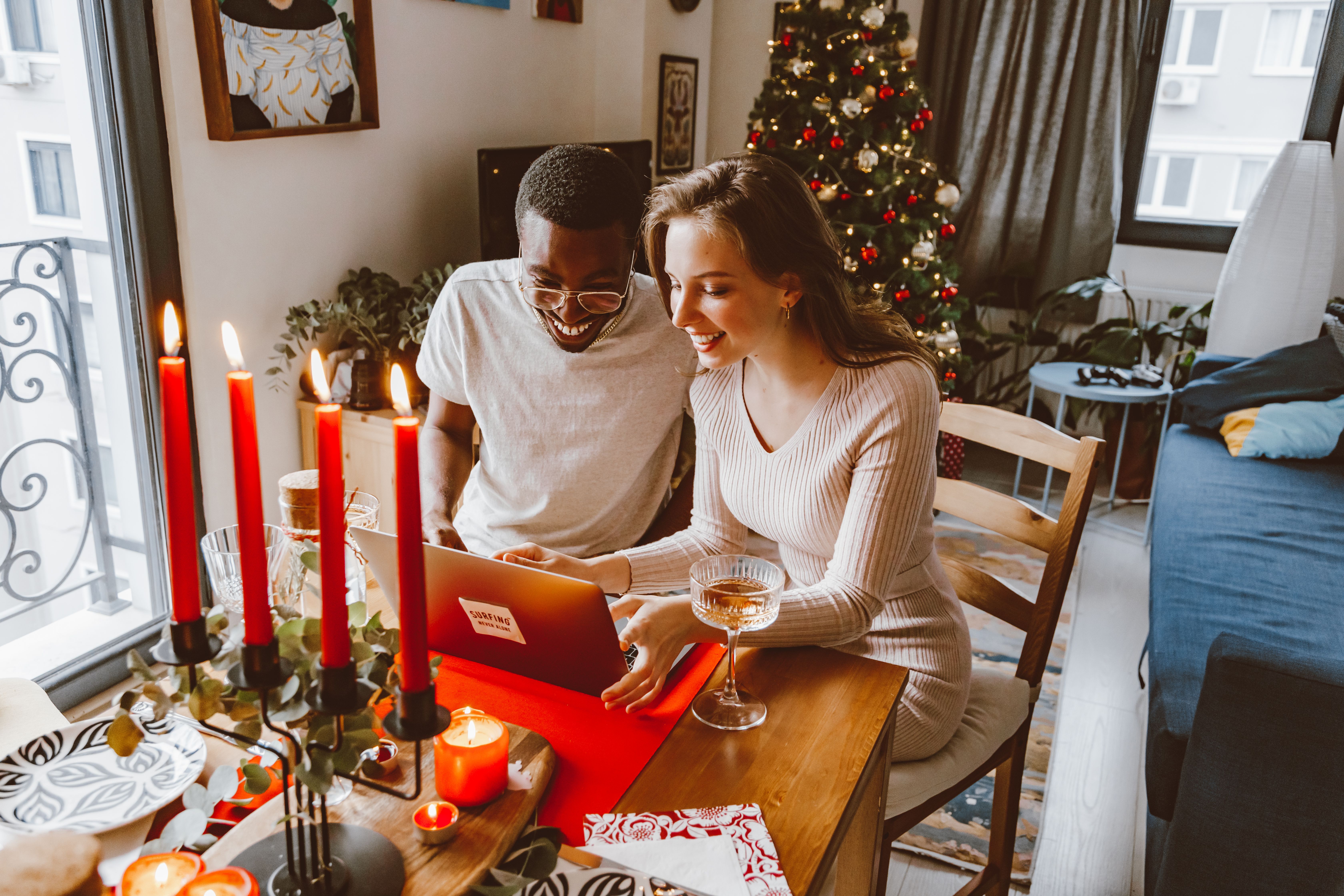 Smiling interracial couple using laptop at festive dinner table indoors, surrounded by Christmas decor.