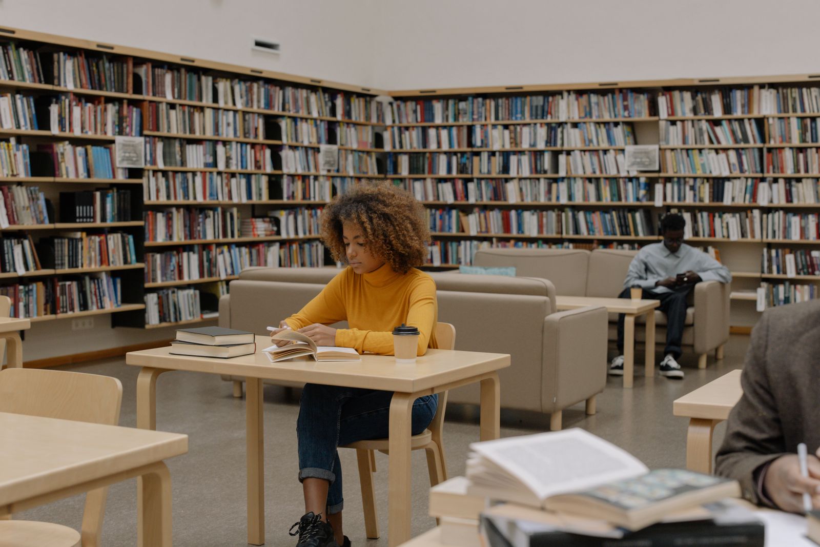 Étudiante lisant un livre à une table de bibliothèque, entourée de rayonnages remplis d'ouvrages.