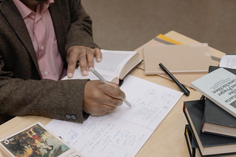 Adult learner writing notes in a notebook with reference books open on the desk