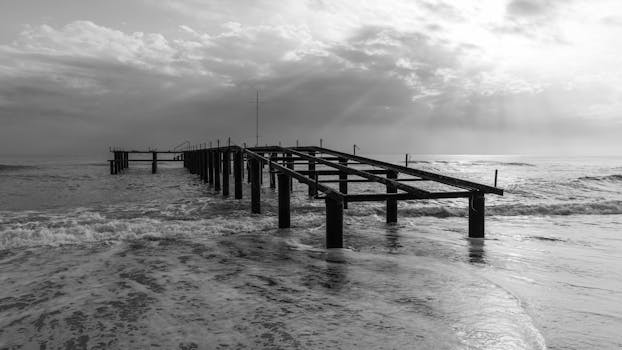 Monochrome image of a dramatic pier extending into the ocean under a cloudy sky.