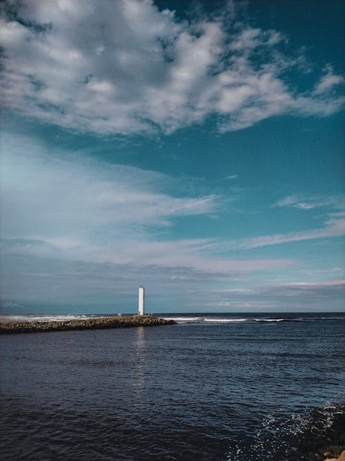 Picturesque scenery of endless ocean and rocky coast with lighthouse on top under cloudy blue sky