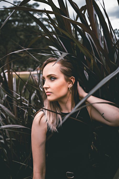 Young contemplative female in black outfit touching hair while looking away under exotic plant in daylight