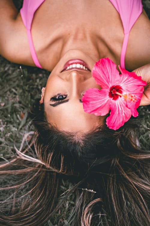 From above of young content woman with toothy smile and blossoming rose mallow looking at camera while lying on grass