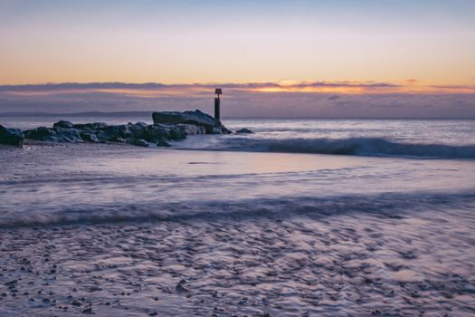 Tranquil scene of sunrise over Bournemouth beach with crashing waves and rocky shore.