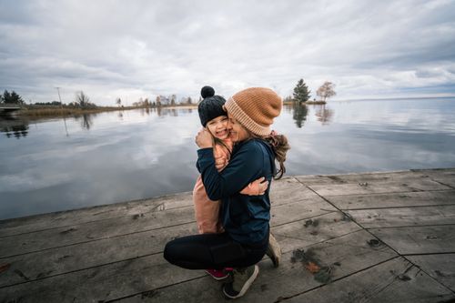 Free A heartwarming moment of a mother and daughter embracing on a lakeside dock under a cloudy sky. Stock Photo
