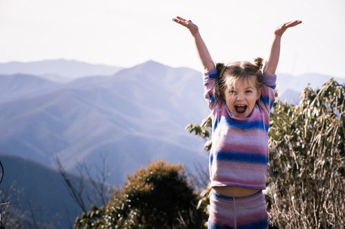Free A cheerful young girl raises her hands in excitement with a scenic mountain range in the background. Stock Photo
