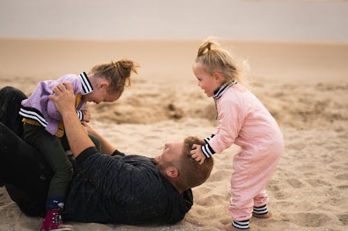 Man in Black T-shirt Carrying Baby in Pink Long Sleeve Shirt on Brown Sand during