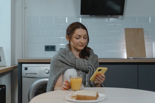 A woman sitting in a modern kitchen, wrapped in a blanket, using her smartphone while having breakfast.