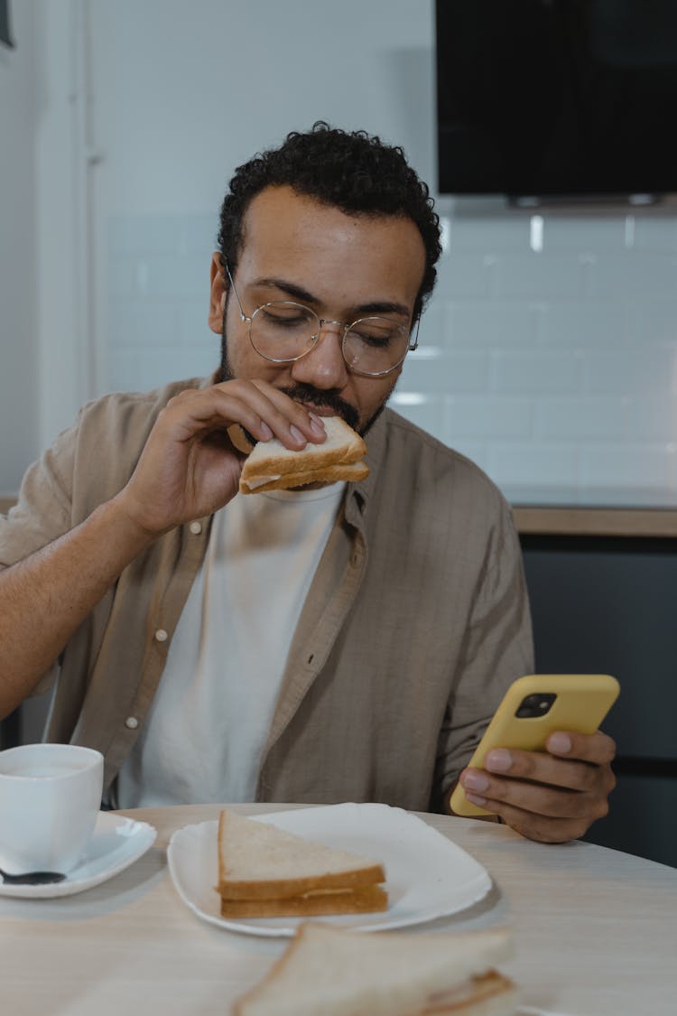 Man In Brown Button Up Shirt Eating Sandwich While Using Yellow Smartphone