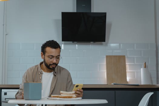 Man with glasses using smartphone at home kitchen table, enjoying meal.