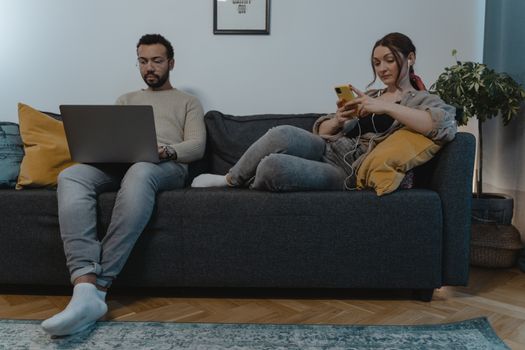 A man and woman relaxing indoors on a sofa, using a laptop and smartphone.
