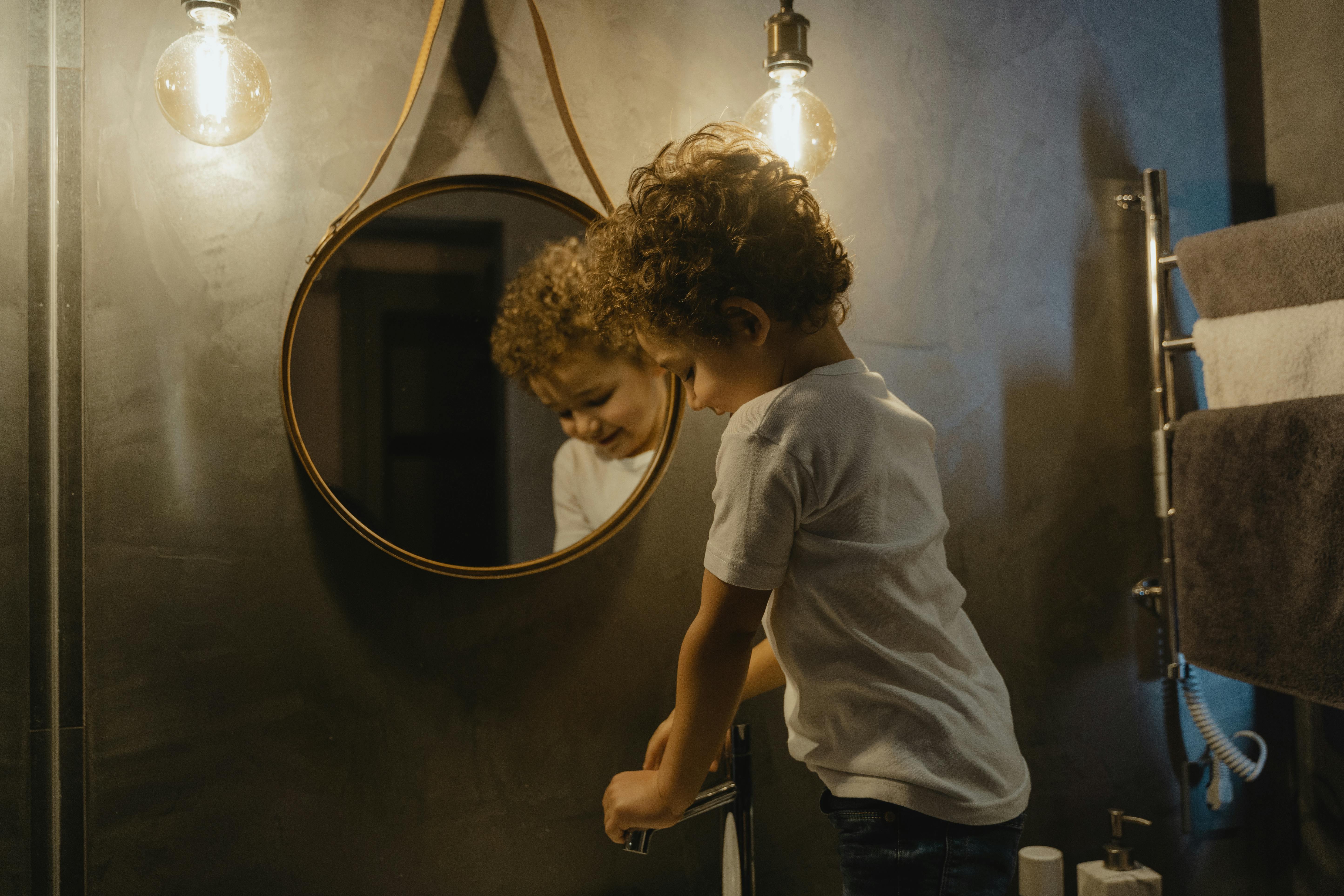 Young boy with curly hair washes hands in bathroom, reflected in mirror.