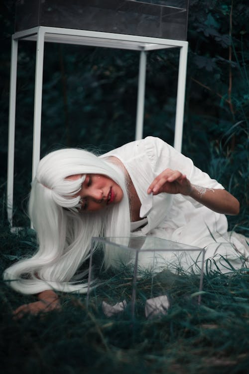 Mysterious Asian female in white wig and white dress sitting on meadow placing origami boats in glass box in summer day