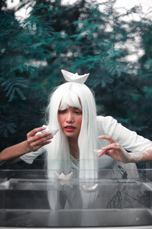 Fantasy ethnic woman in white wig and dress placing paper boats on water of aquarium in mysterious woods