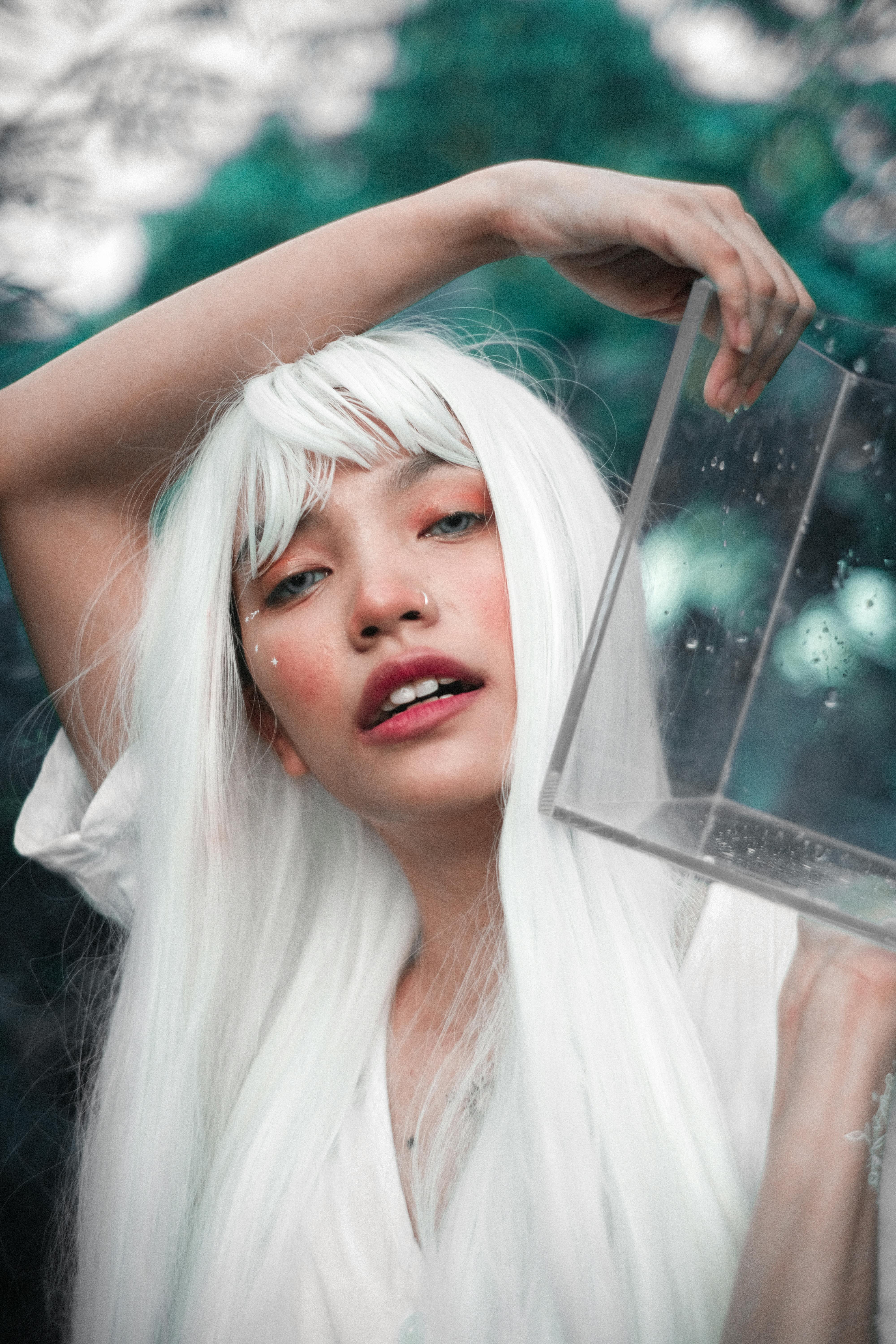 Tender ethnic woman in white dress and white wig carrying transparent aquarium against summer trees looking at camera