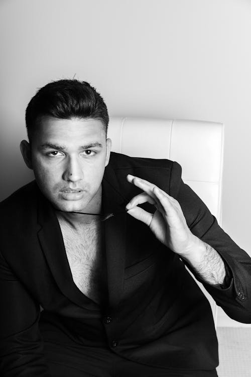 Black and white of handsome stylish male in black suit sitting on white leather chair and looking at camera