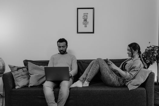 A diverse couple using gadgets on a sofa, focused on their tasks in a black-and-white setting.