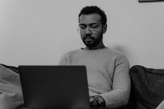 Serious bearded man in sweater working remotely from home using laptop, captured in black and white.