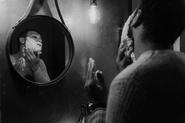 A Man Putting Shaving Foam On His Face While Looking At The Round Mirror