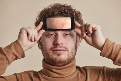 Free Man with curly hair and facial hair holds smartphone on forehead in neutral studio background. Stock Photo