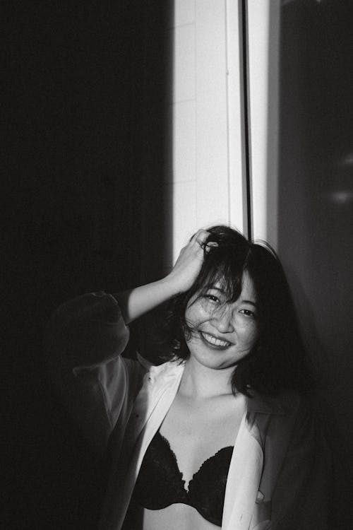 Black and white of smiling young Asian female in bra and shirt leaning on wall in dark room with sunlight and looking at camera while touching hair with hand